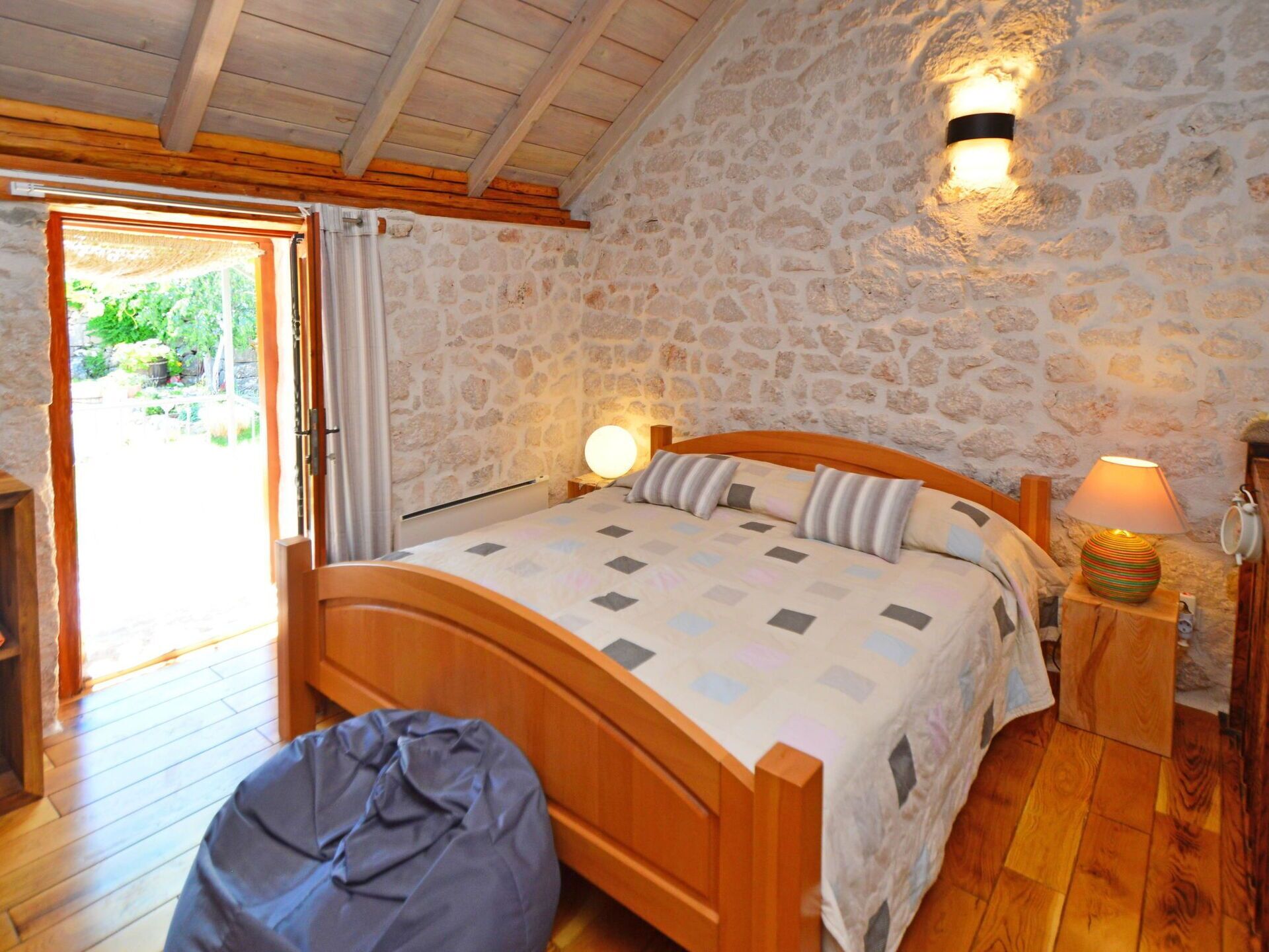 A bedroom with a stone wall, wooden ceiling beams, a bed with a patterned bedspread, two bedside lamps, and a small round table with a lamp.