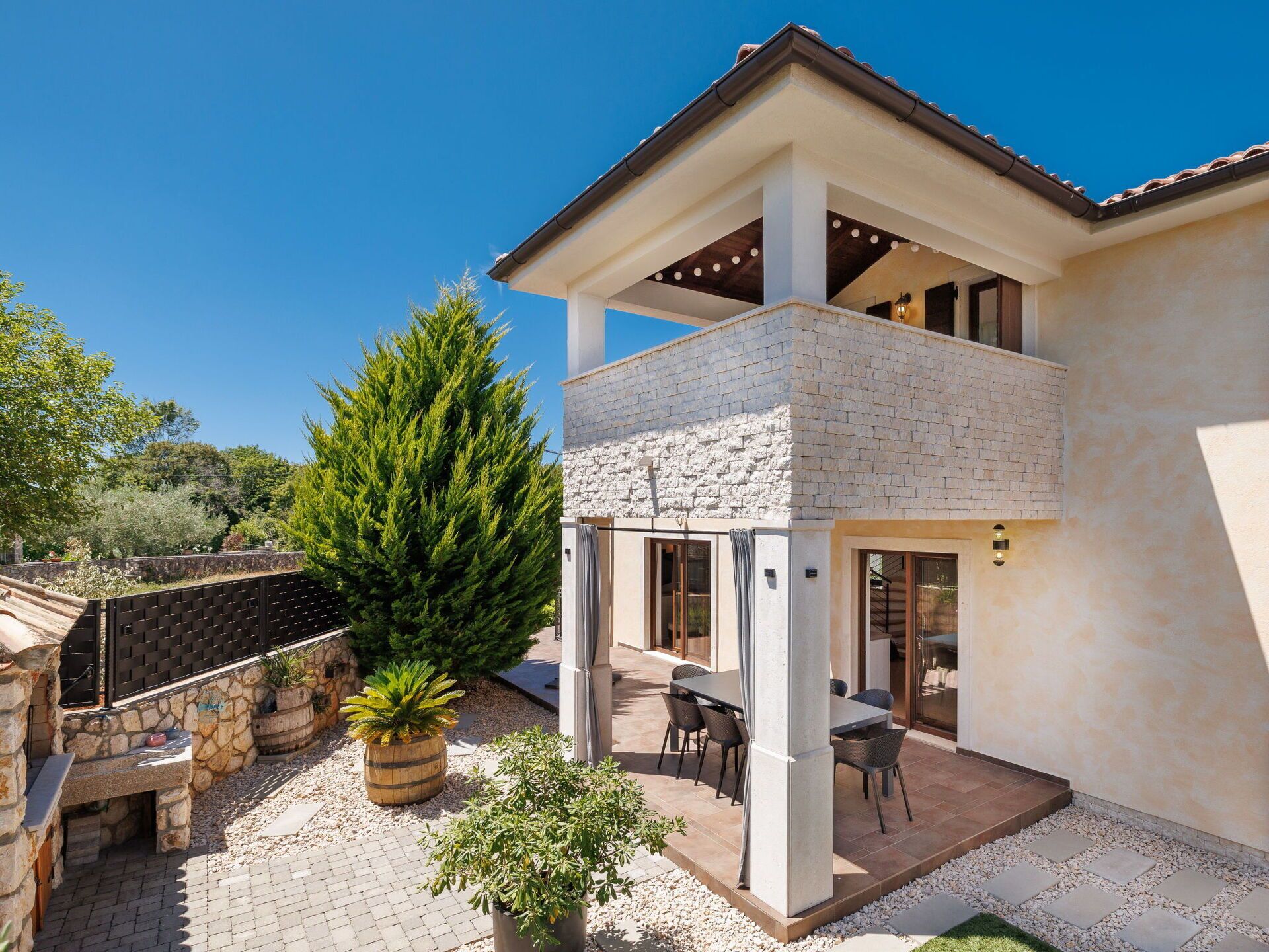 Uma casa de dois andares com fachada de pedra, varanda com assento externo e área de jardim com plantas em vasos.