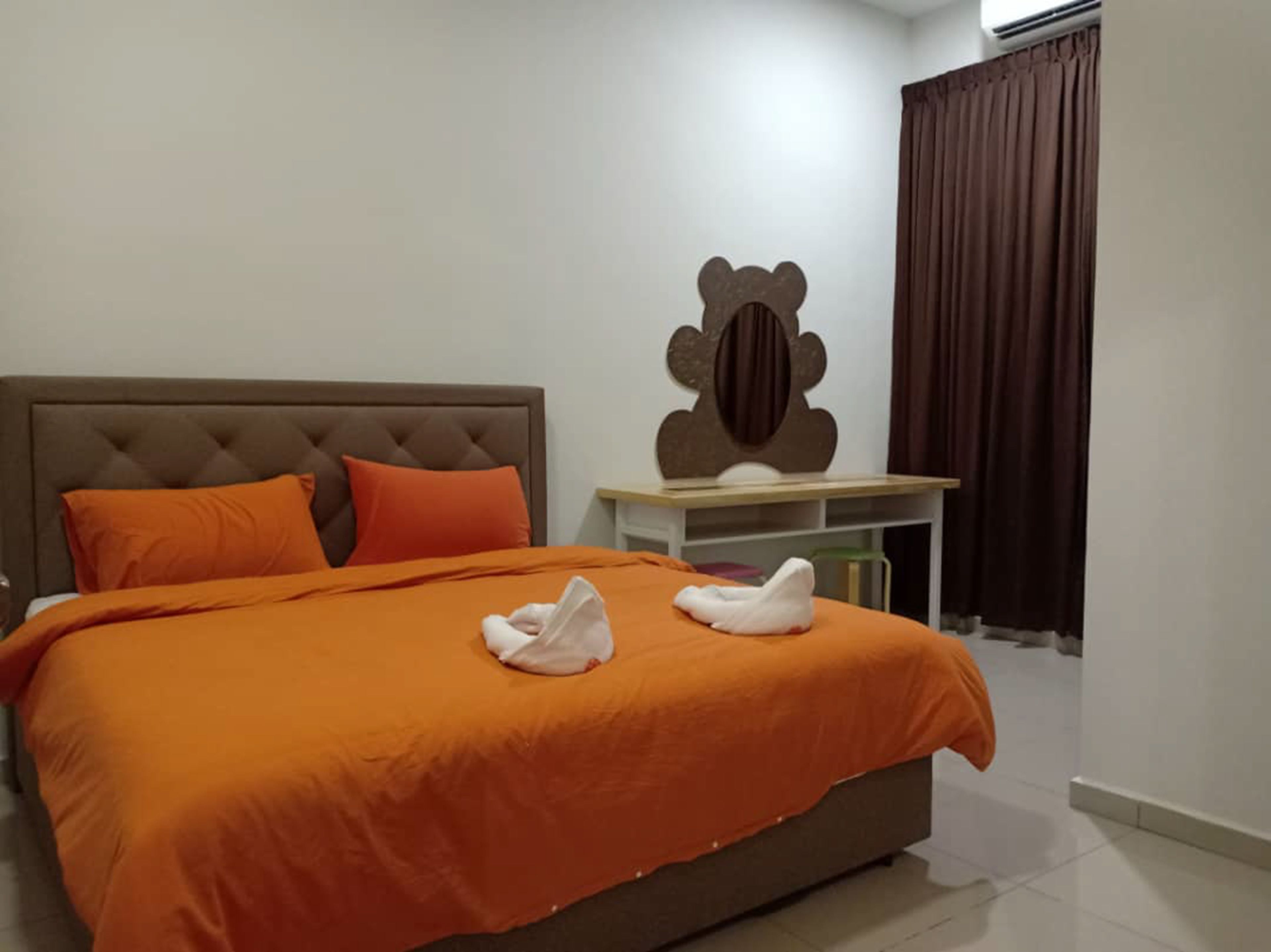 A bedroom with a large bed, orange bedding, a tufted headboard, a wooden side table, a mirror, and a window with curtains.