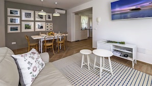 Una sala moderna con sofá, un comedor con mesa y sillas, una cocina al fondo y un cuadro grande en la pared.