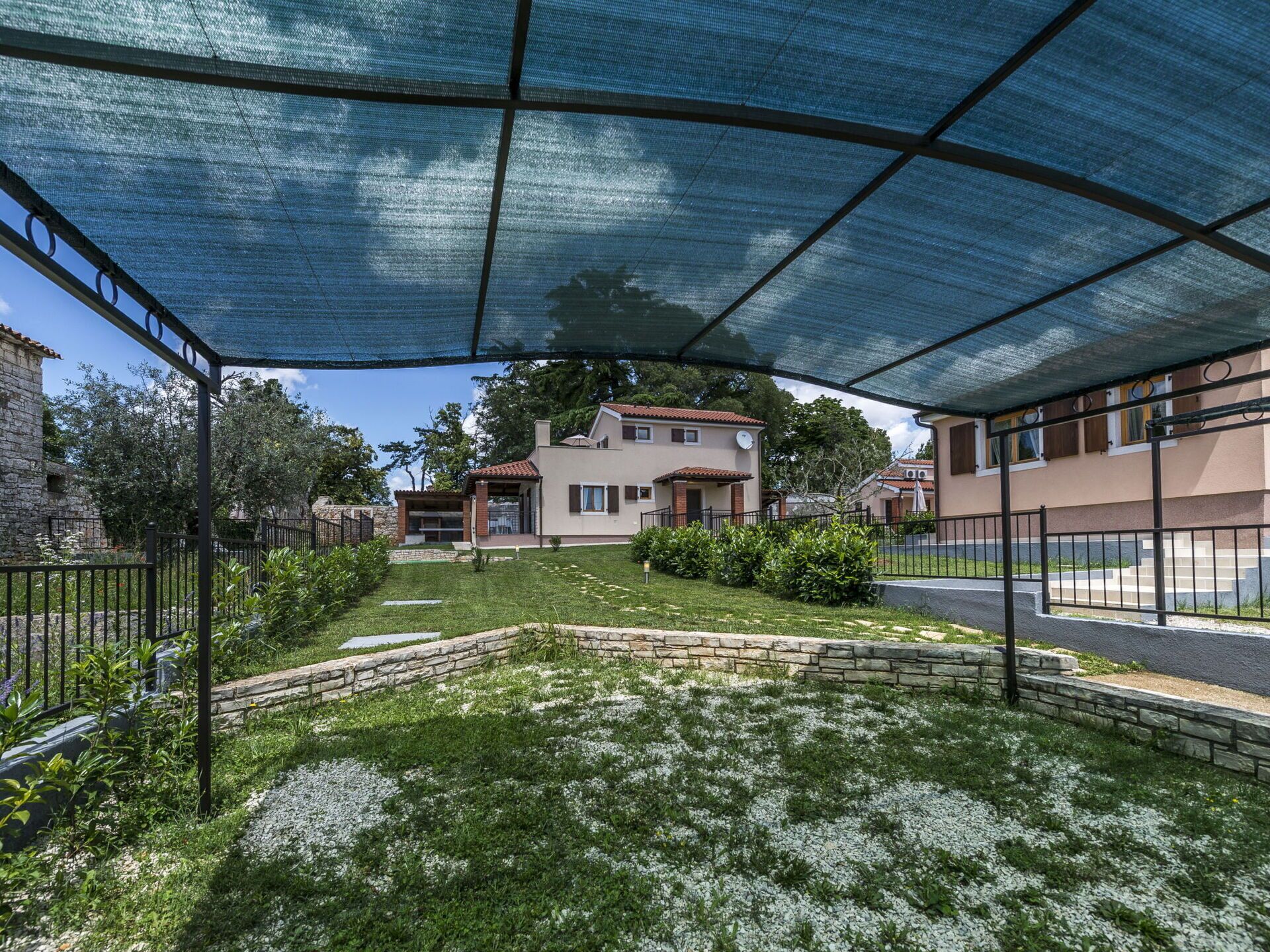 A residential area with a house under a shaded structure, surrounded by greenery and stone walls.