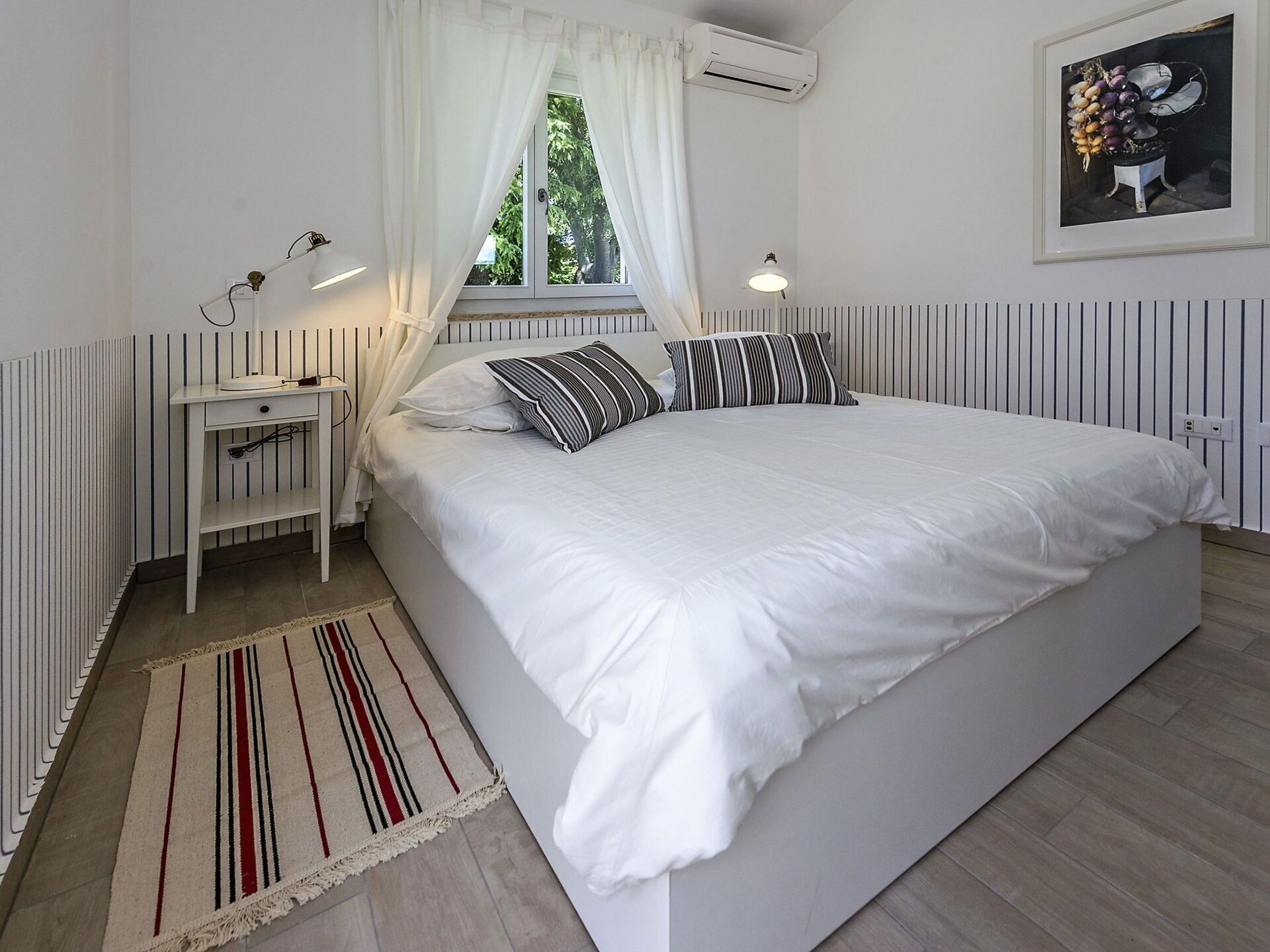 A bedroom with a large bed, a bedside table, a lamp, a window with curtains, an air conditioning unit, and a framed picture on the wall.