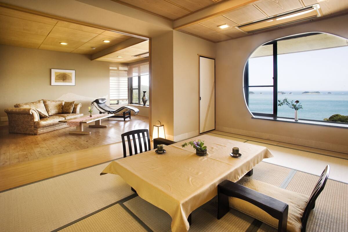 A traditional Japanese-style room with tatami flooring, a low table with cushions, a sofa, and a view of the sea through a large window.