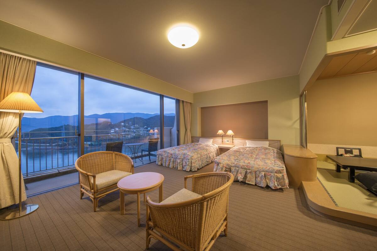 A hotel room with a balcony, two beds with floral bedding, wicker furniture, and a view of the city and mountains.