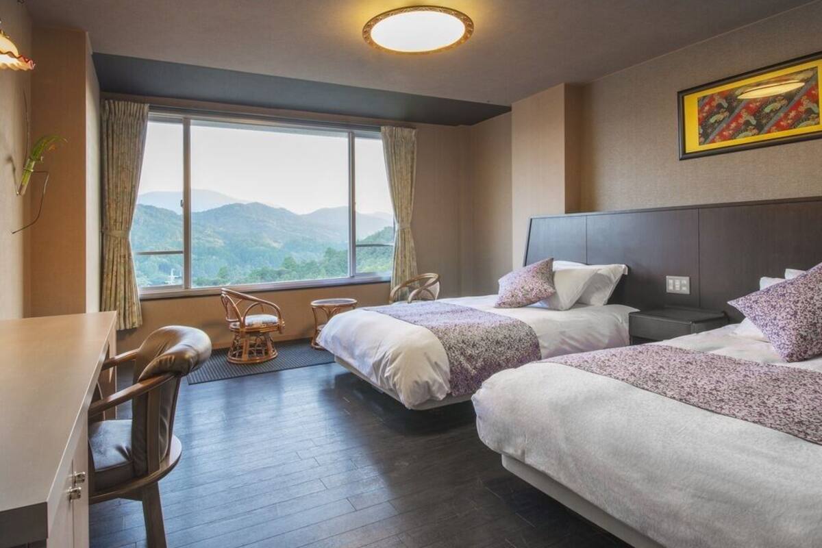 A hotel room with two beds, a desk, a chair, a window with a mountain view, and a ceiling light.
