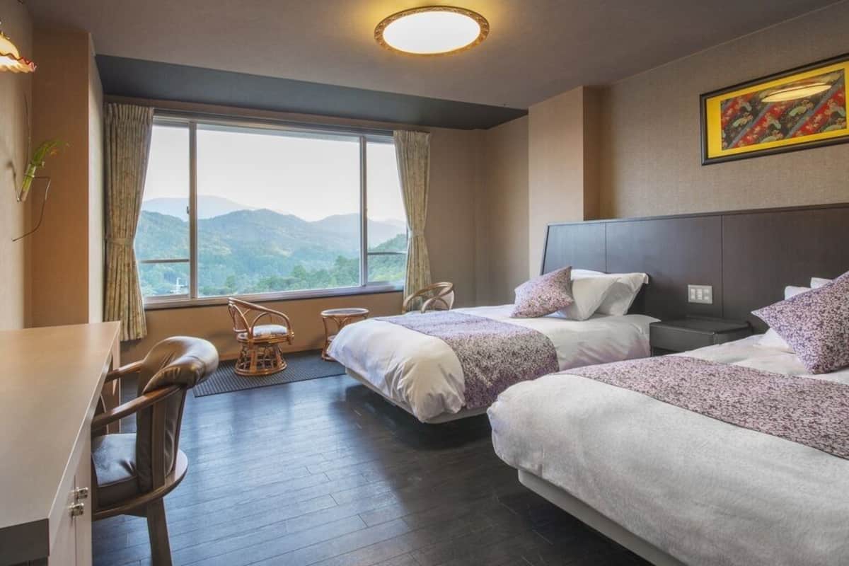 A hotel room with two beds, a desk, a chair, a window with a mountain view, and a ceiling light.
