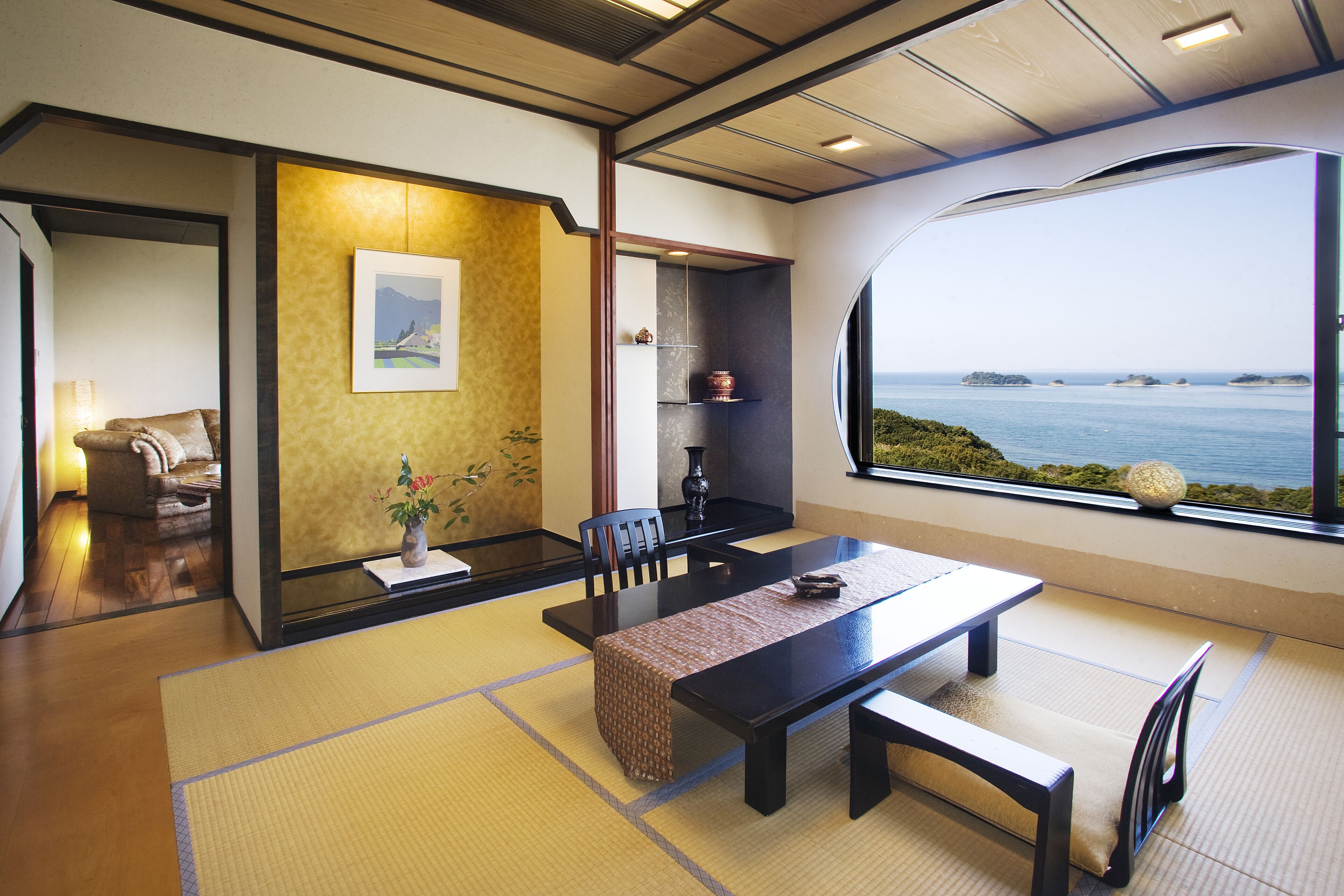 Une pièce de style japonais traditionnel, avec un sol recouvert de tatamis, une table basse entourée de bancs, un canapé et une vue sur la mer par une grande fenêtre.