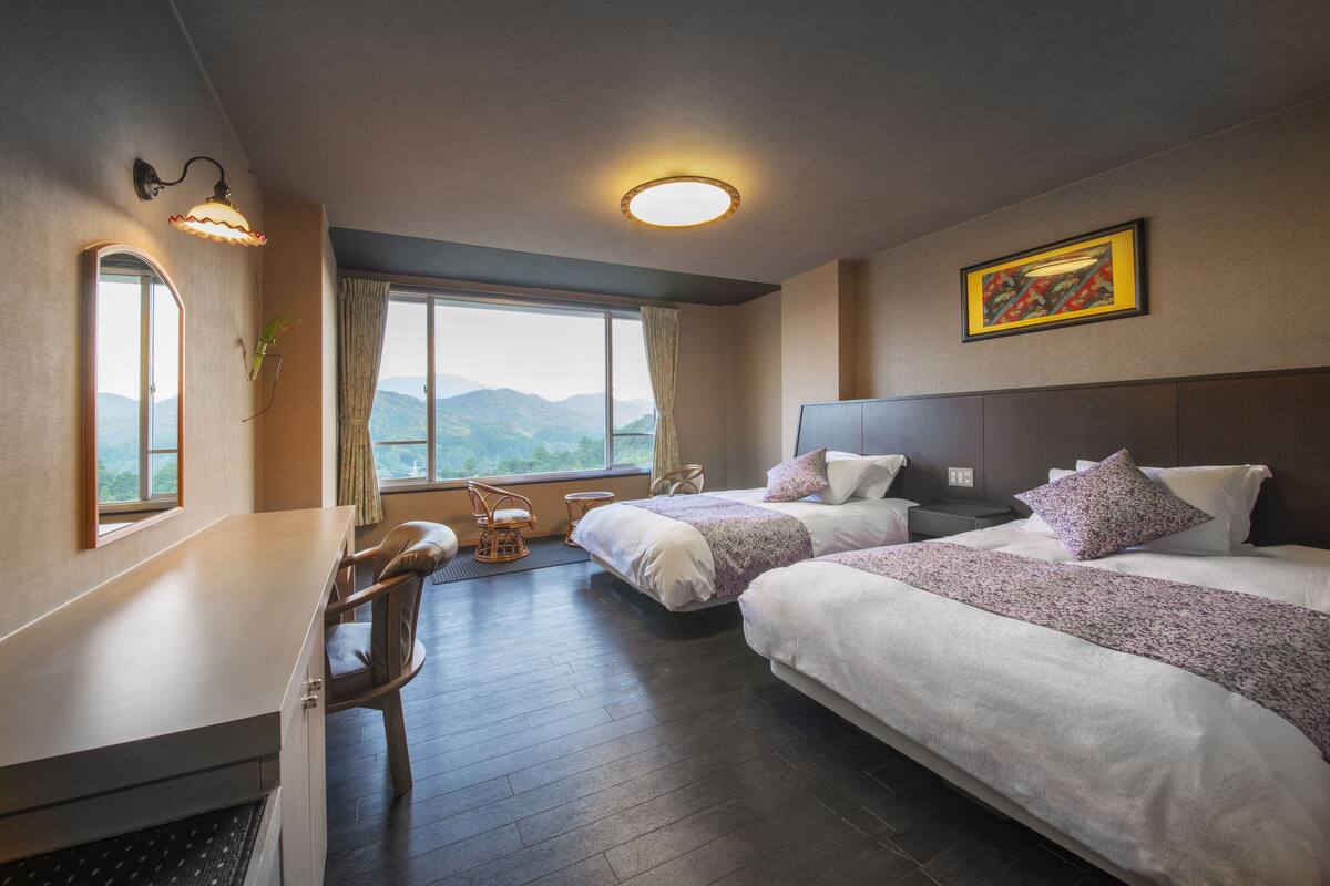 A hotel room with two beds, a desk, a chair, a window with a view of mountains, and a painting on the wall.