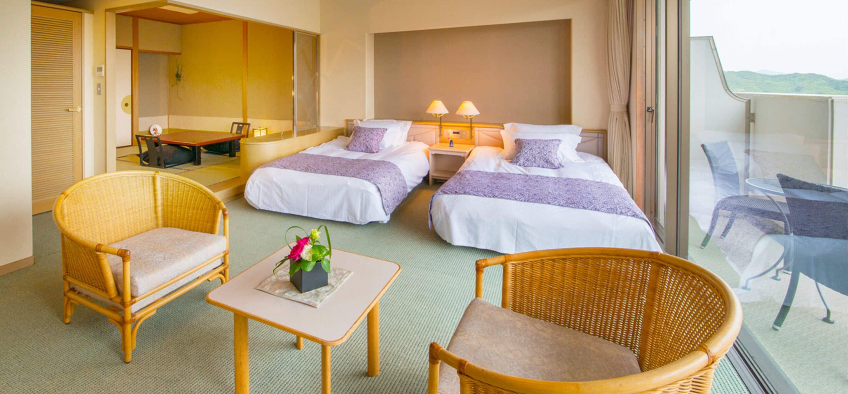 A hotel room with two beds, a dining table, a small table with a flower, two wicker chairs, and a balcony with a view.
