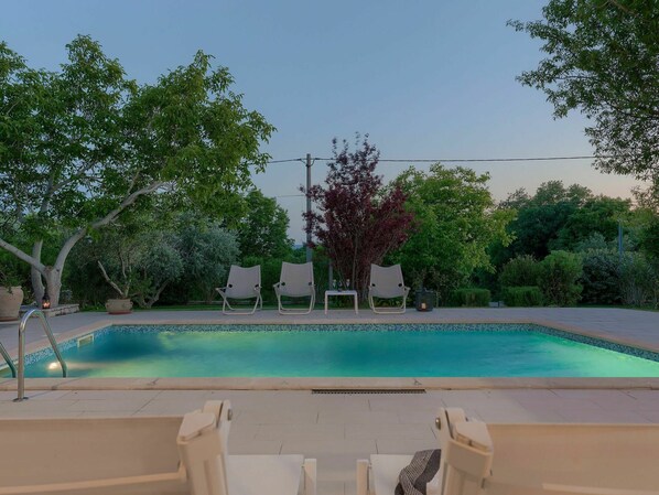 Ein rechteckiger Pool mit einer gefliesten Kante, umgeben von einer gepflasterten Liegefläche. Mehrere Liegestühle und ein kleiner Tisch mit einer Lampe sind vorhanden.