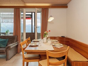 A dining area with a wooden table set for four, chairs with cushioned seats, a vase with flowers, and a view of mountains through a glass door.