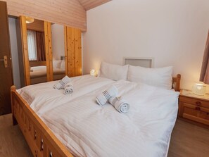 Un lit double avec du linge de lit blanc et deux serviettes enroulées, une tête de lit en bois, une table de chevet avec une lampe et une armoire en bois avec un miroir.