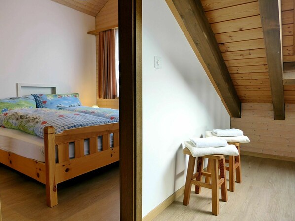 Un lit en bois avec une couvre-lit à motifs floraux, une tête de lit en bois et une table de chevet avec une lampe. On y trouve également un tabouret en bois sur lequel est posée une serviette.