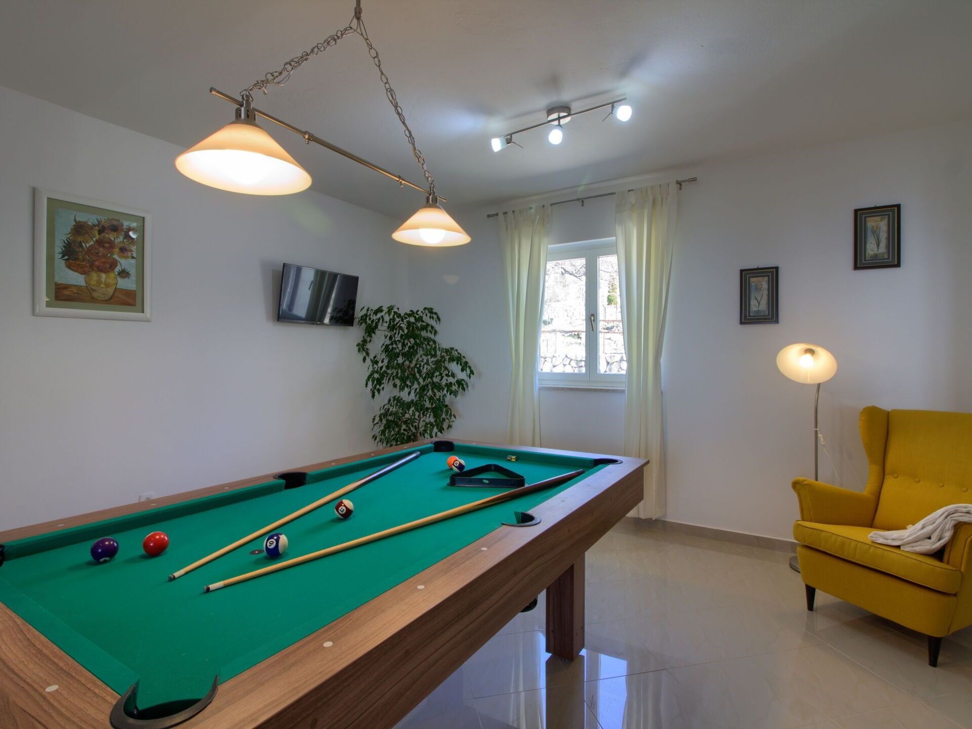 Une table de billard avec des billes, un fauteuil jaune, une plante et un téléviseur accroché au mur.