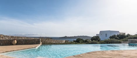 A swimming pool with a stone deck, a stone wall, and a view of the sea and distant hills.