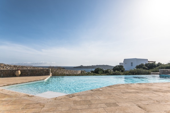 A swimming pool with a stone deck, a stone wall, and a view of the sea and distant hills.