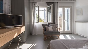 Un dormitorio moderno con un escritorio de madera, televisión, una butaca cómoda con cojines, cama y un amplio ventanal con cortinas.