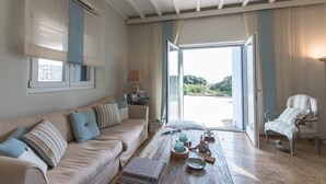 Amplia sala de estar con un sofá color beige, una mesa de centro de madera y un sillón a rayas. Un ventanal ofrece vistas a zonas verdes.