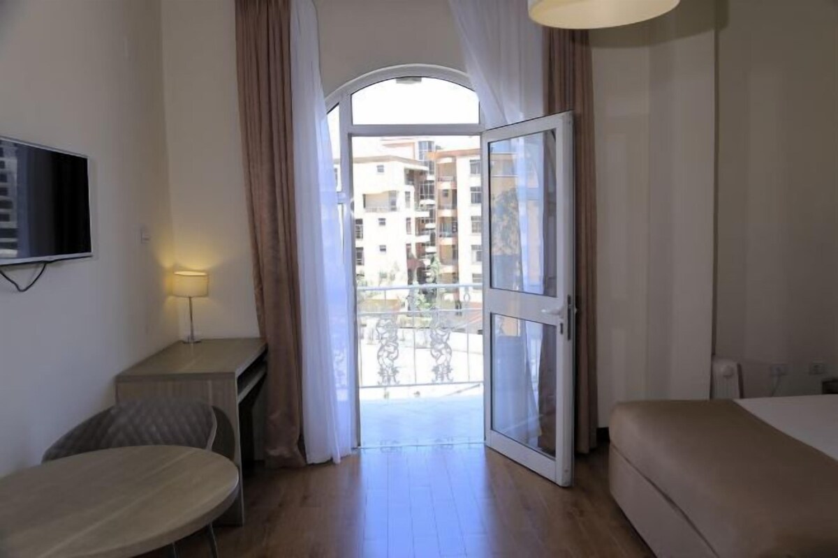 A hotel room with a bed, a small table with a lamp, a TV mounted on the wall, and a balcony with a view of other buildings.