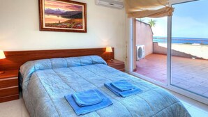 A bedroom with a bed, a nightstand, a lamp, a painting on the wall, and a view of the beach through a sliding door.