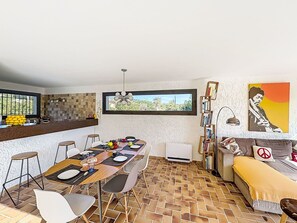 Un comedor con una mesa preparada para cuatro, una barra de cocina con fruta, un ventanal con vistas y una estantería llena de libros.