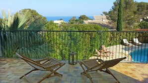 En balkon med to træloungestole, et lille bord og udsigt til en swimmingpool og havet.