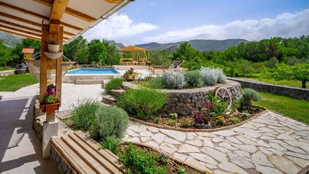 Un espace piscine extérieur agrémenté d’une pergola en bois, d’un chemin en pierre et d’un jardin où poussent différentes espèces de plantes, ainsi que d’un kiosque.