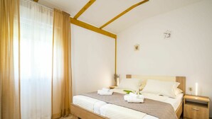 Une chambre à coucher bien rangée, avec un lit en bois, du linge de lit blanc, une table de chevet avec une lampe et une fenêtre ornée de rideaux légers.
