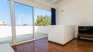 Ein geräumiger Balkon mit einer Schiebeglastür, einem Flachbildfernseher auf einem Standfuß und Blick auf ein Gebäude und Bäume.