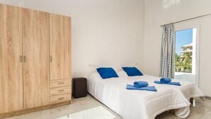 Ein Schlafzimmer mit einem großen Holzschrank, einem Bett mit weißer Bettwäsche und blauen Kissen sowie Blick auf ein Gebäude durch ein Fenster.