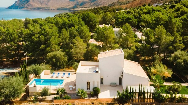 Eine moderne Villa mit Swimmingpool, umgeben von üppiger Vegetation mit Blick auf eine gebirgige Küstenlinie.