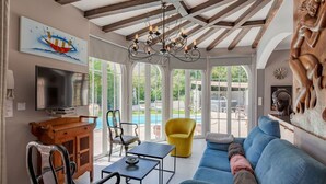 Amplia sala de estar con un sofá azul, una mesa de centro de madera y un televisor. Hay un mueble de madera, un sillón amarillo y un amplio ventanal con vistas a una piscina.