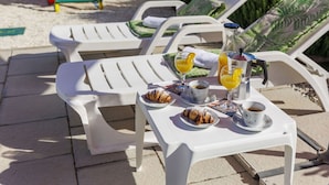 A sunlit outdoor seating area with white lounge chairs, a table with croissants, coffee, and orange juice.