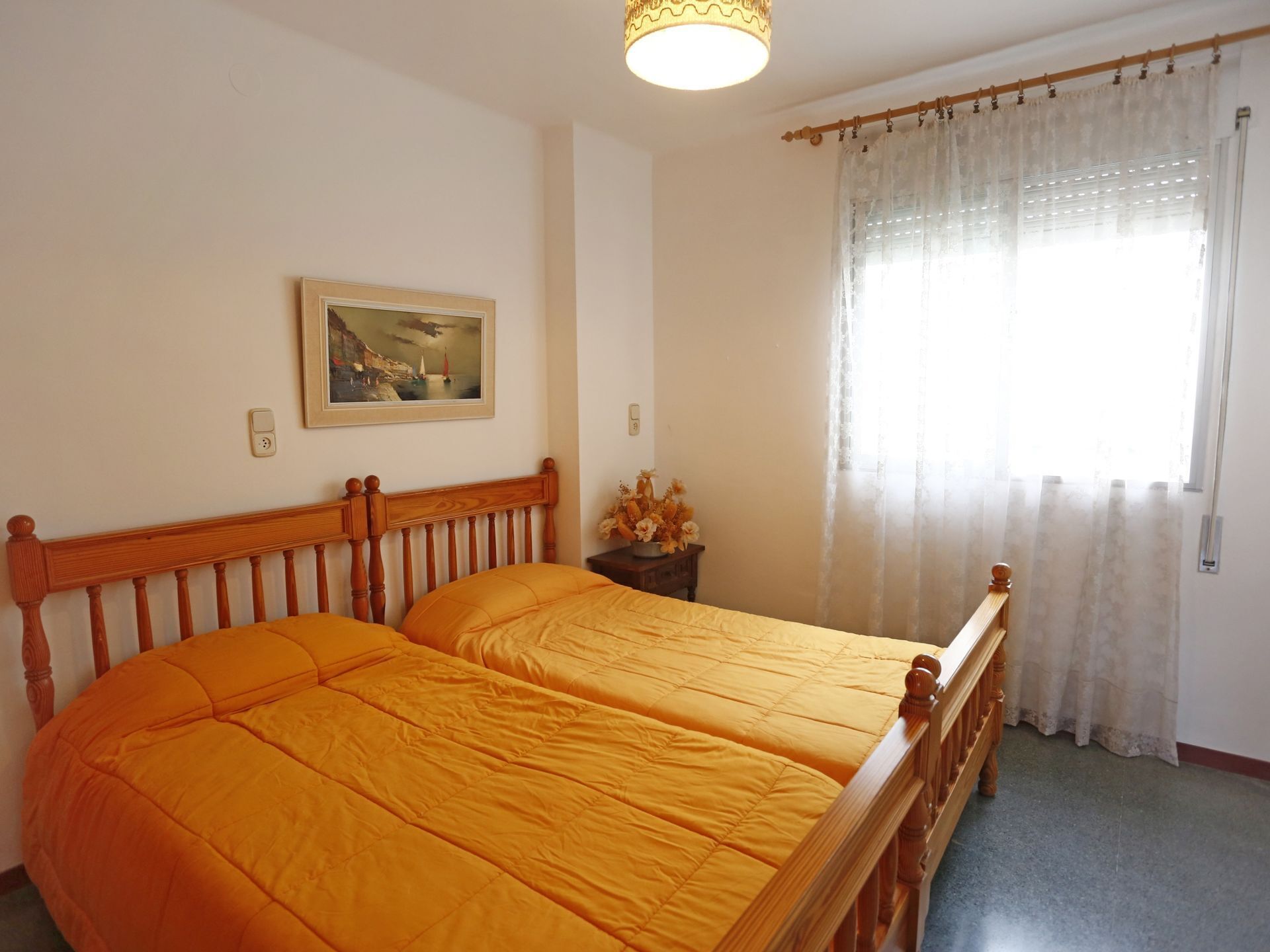 Ein Schlafzimmer mit Doppelbett, einem Bild an der Wand, einem Nachttisch mit Blumenvase und einem Fenster mit durchsichtigen Vorhängen.