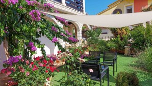 Ein Garten mit verschiedenen Blumen und Pflanzen, eine Sitzgelegenheit mit Stühlen sowie ein Gebäude mit weißer Fassade und Bögen.