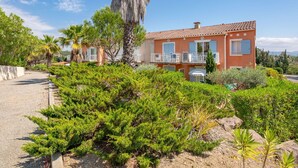 Un edificio de dos pisos con fachada roja, balcones y un jardín bien cuidado con diversos arbustos y una palmera.