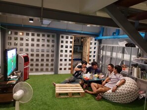 A group of people sitting on a green artificial turf floor, with a television and fan in the background. - Hybrit Hostel & Cafe (Hat Yai)