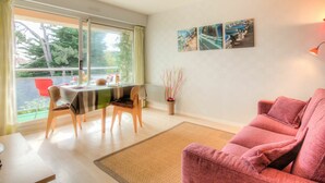 A modern living room with a sofa, a dining table with chairs, and a balcony with a view of trees and buildings. - Cosy apartment for 2 guests with WIFI, TV, balcony and pets allowed (Dinard)