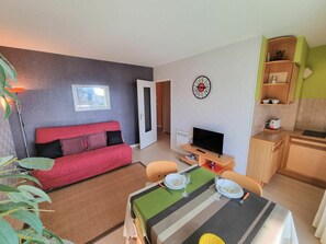 Un espace de vie compact comprenant un canapé rouge, une table à manger dressée avec des assiettes et des verres, un téléviseur et un coin cuisine avec une horloge au mur.