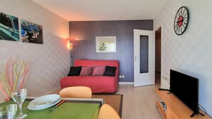 A compact living space with a dining area, a red sofa, a television, and a clock on the wall. - Cosy apartment for 2 guests with WIFI, TV, balcony and pets allowed (Dinard)