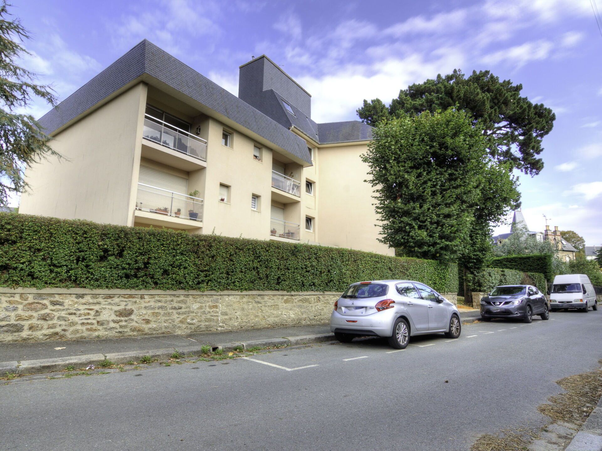 Un edificio residencial de varios pisos con balcones, coches aparcados y un muro de piedra.