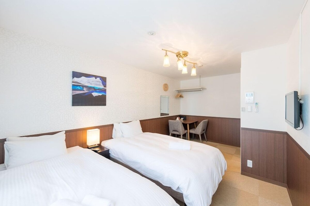 A hotel room with two beds, a desk, and a chair. There is a painting on the wall and a ceiling light fixture.