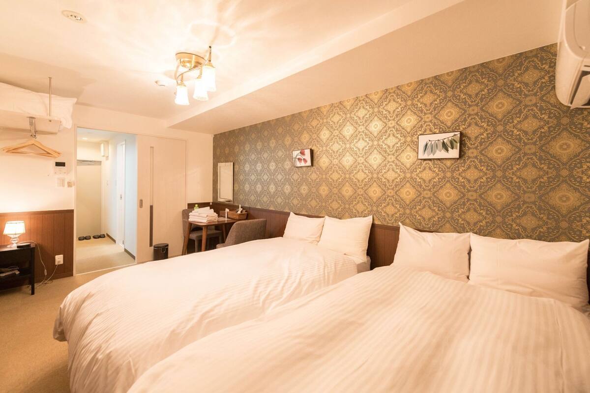 A hotel room with two beds, a desk, a chair, a hanging coat rack, and a patterned wallpaper.