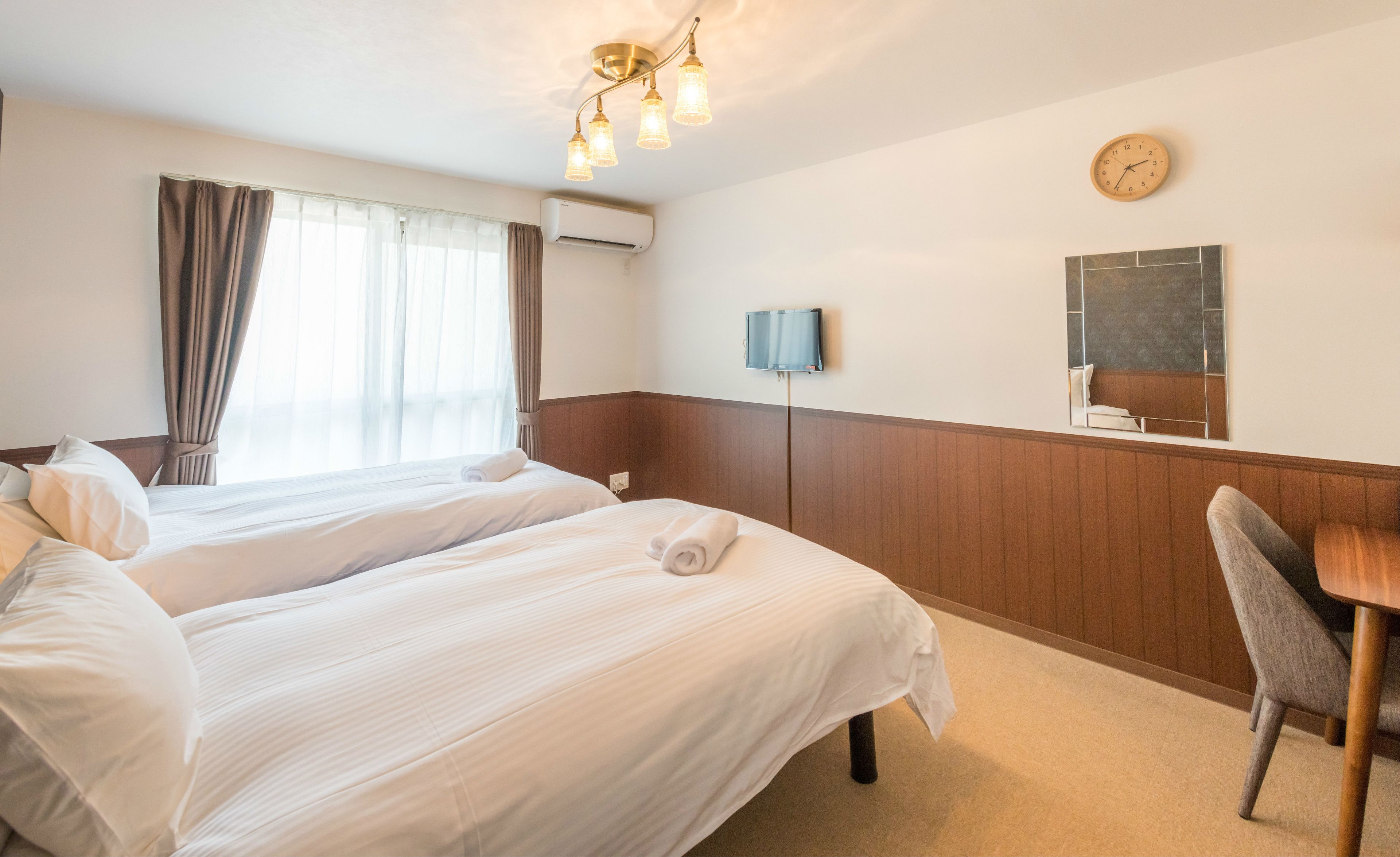A hotel room with two beds, a desk, a television, a clock, and a window with curtains.