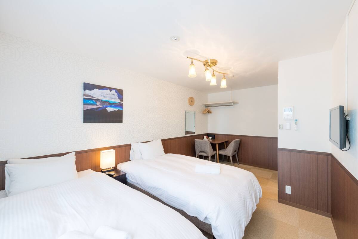 A hotel room with two beds, a desk, and a chair. There is a painting on the wall and a clock.