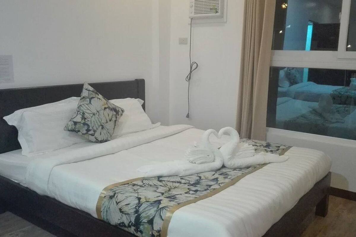 A neatly made bed with white linens and a floral-patterned throw, a dark headboard, and a window with a view of a building.