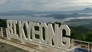 Um grande letreiro branco com o texto "BEKMEONG" ao fundo de uma paisagem montanhosa e um corpo de água.