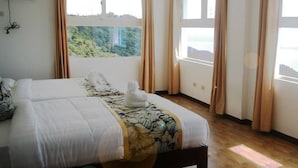 Quarto de hotel com uma cama grande, duas janelas com cortinas, vista para o verde lá fora e piso de madeira.
