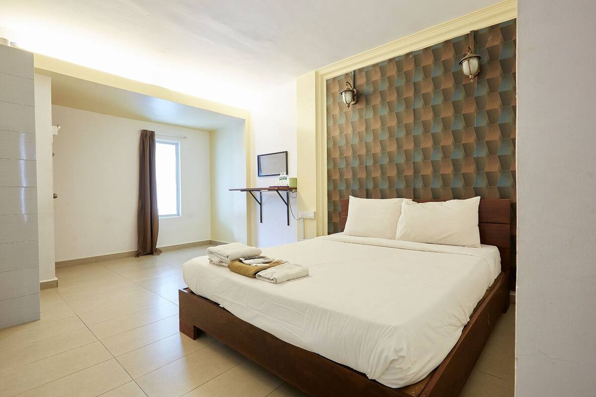 A hotel room with a large bed, a wooden headboard, a flat-screen TV, and a small table with a lamp.
