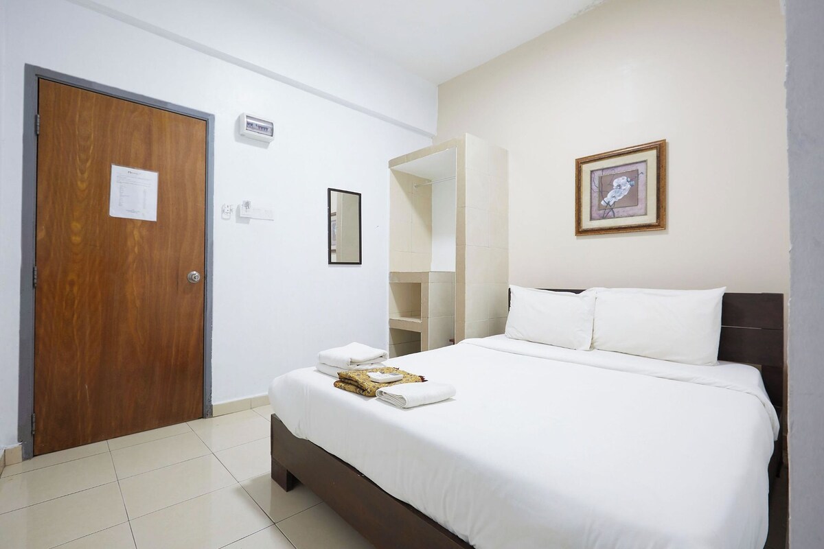 A hotel room with a wooden door, a bed with white linens, a framed picture on the wall, and a small shelf with toiletries.